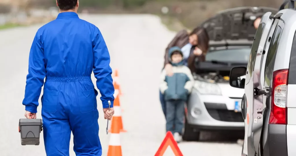 A person in blue jumpsuit standing next to a car with a child in the background